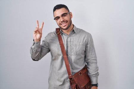Foto de Un joven hispano con una maleta sonriendo mirando a la cámara mostrando los dedos haciendo señal de victoria. número dos. - Imagen libre de derechos