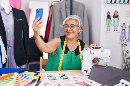 Téléchargez les photos : Femme aux cheveux gris moyen âge tailleur souriant confiant faire selfie par smartphone au magasin de tailleur - en image libre de droit