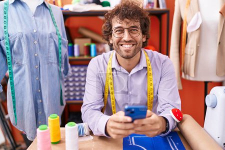 Téléchargez les photos : Jeune homme hispanique tailleur souriant confiant en utilisant smartphone à l'atelier - en image libre de droit
