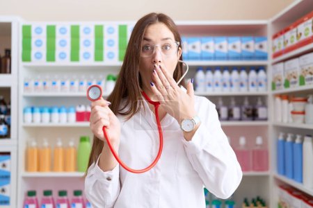 Téléchargez les photos : Belle femme brune travaillant à la pharmacie pharmacie tenant stéthoscope couvrant la bouche à la main, choquée et effrayée par l'erreur. expression surprise - en image libre de droit