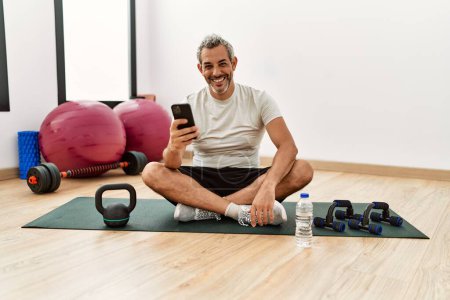 Foto de Hombre de pelo gris de mediana edad sentado en una esterilla de yoga usando un teléfono inteligente en el centro deportivo - Imagen libre de derechos