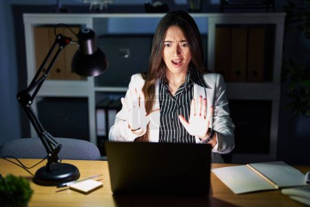 Téléchargez les photos : Jeune femme brune travaillant au bureau la nuit avec un ordinateur portable faisant geste d'arrêt avec les mains paumes de main, expression de colère et de frustration - en image libre de droit
