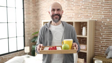 Photo for Young bald man holding breakfast tray standing at bedroom - Royalty Free Image