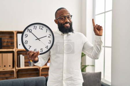 Foto de African american man holding big clock at the office smiling with an idea or question pointing finger with happy face, number one - Imagen libre de derechos