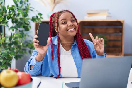 Téléchargez les photos : Femme afro-américaine avec des cheveux tressés tenant smartphone montrant écran vide surpris par une idée ou une question pointant du doigt avec un visage heureux, numéro un - en image libre de droit