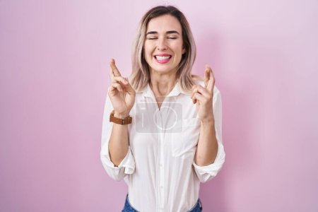 Téléchargez les photos : Young beautiful woman standing over pink background gesturing finger crossed smiling with hope and eyes closed. luck and superstitious concept. - en image libre de droit