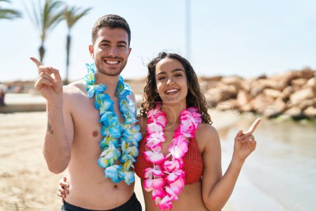 Téléchargez les photos : Jeune couple hispanique portant collier hawaïen à la plage souriant heureux pointant avec la main et le doigt sur le côté - en image libre de droit