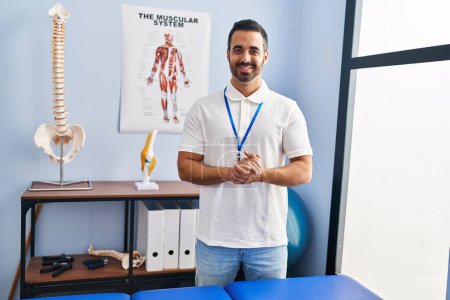 Téléchargez les photos : Jeune homme hispanique physiothérapeute souriant confiance debout à la clinique de réadaptation - en image libre de droit