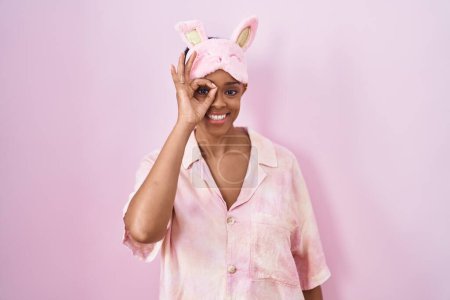 Foto de Mujer afroamericana con máscara de sueño y pijama sonriendo feliz haciendo signo de bien con la mano en el ojo mirando a través de los dedos - Imagen libre de derechos