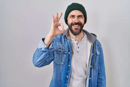 Téléchargez les photos : Jeune homme hispanique avec des tatouages portant un bonnet de laine souriant positif faisant signe ok avec la main et les doigts. expression réussie. - en image libre de droit