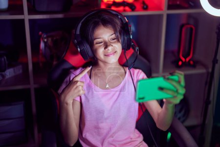 Foto de Chica joven jugando videojuegos con teléfono inteligente sonriendo feliz señalando con la mano y el dedo - Imagen libre de derechos