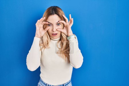 Foto de Young caucasian woman standing over blue background trying to open eyes with fingers, sleepy and tired for morning fatigue - Imagen libre de derechos