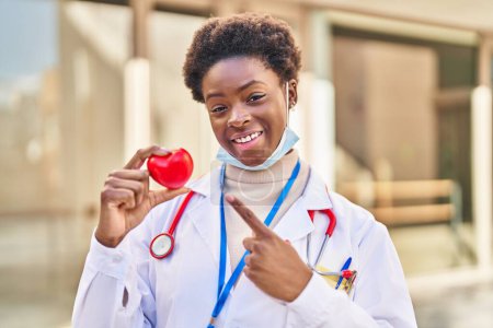 Téléchargez les photos : Femme afro-américaine portant l'uniforme de médecin tenant le cœur souriant heureux pointant avec la main et le doigt - en image libre de droit