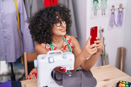 Téléchargez les photos : Jeune femme du Moyen-Orient tailleur à l'aide d'une machine à coudre faire selfie par smartphone au magasin de tailleur - en image libre de droit