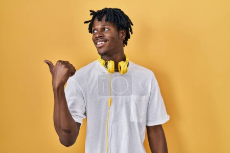 Téléchargez les photos : Jeune homme africain avec dreadlocks debout sur fond jaune souriant avec le visage heureux regardant et pointant vers le côté avec le pouce vers le haut. - en image libre de droit