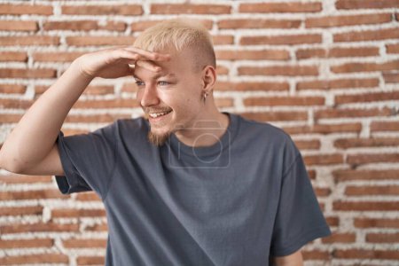 Photo for Young caucasian man standing over bricks wall very happy and smiling looking far away with hand over head. searching concept. - Royalty Free Image