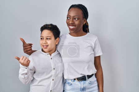 Téléchargez les photos : Jeune mère et son fils debout ensemble sur fond blanc pointant vers le dos derrière avec la main et les pouces levés, souriant confiant - en image libre de droit