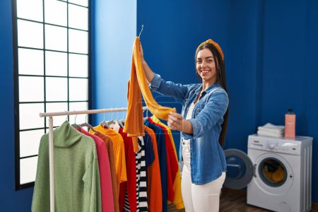 Téléchargez les photos : Young beautiful hispanic woman holding clothes on rack at laundry room - en image libre de droit