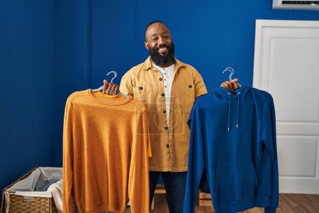 Photo for African american man holding clean clothes on hangers at laundry room smiling with a happy and cool smile on face. showing teeth. - Royalty Free Image