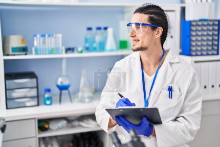 Téléchargez les photos : Jeune homme portant uniforme scientifique écrire sur presse-papiers au laboratoire - en image libre de droit