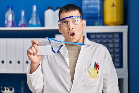 Foto de Hombre árabe joven trabajando en laboratorio científico sosteniendo tarjeta de identificación asustado y sorprendido con la boca abierta para sorpresa, cara de incredulidad - Imagen libre de derechos