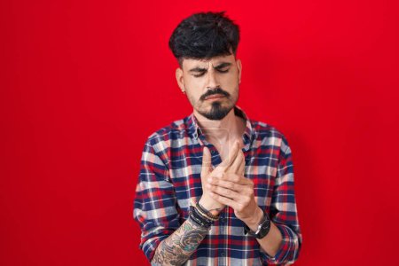 Photo for Young hispanic man with beard standing over red background suffering pain on hands and fingers, arthritis inflammation - Royalty Free Image