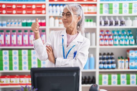 Photo for Middle age woman with tattoos working at pharmacy drugstore looking proud, smiling doing thumbs up gesture to the side - Royalty Free Image