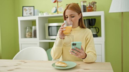 Téléchargez les photos : Jeune femme blonde utilisant un smartphone prenant le petit déjeuner à la maison - en image libre de droit