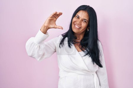 Téléchargez les photos : Femme hispanique mature debout sur fond rose geste souriant et confiant avec la main faisant signe de petite taille avec les doigts regardant et la caméra. concept de mesure. - en image libre de droit
