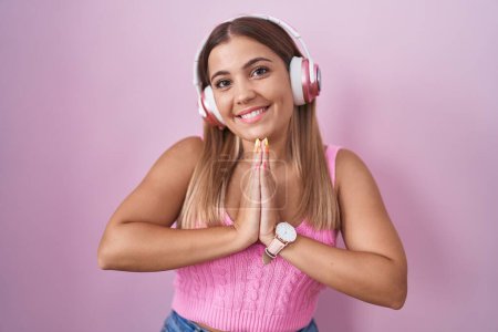Téléchargez les photos : Jeune femme blonde écoutant de la musique à l'aide d'écouteurs priant avec les mains ensemble demandant pardon en souriant confiant. - en image libre de droit
