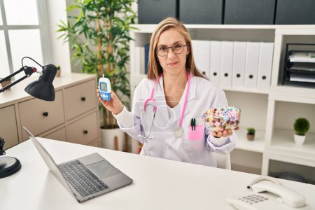 Photo for Young blonde doctor woman holding glucometer and sweets clueless and confused expression. doubt concept. - Royalty Free Image