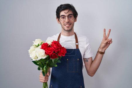 Téléchargez les photos : Jeune homme hispanique tenant un bouquet de roses blanches et rouges souriant avec un clin d'œil heureux à la caméra faisant signe de victoire avec les doigts. numéro deux. - en image libre de droit