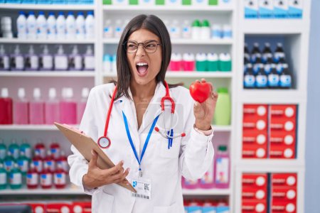 Photo for Brunette young woman working at pharmacy drugstore holding red heart angry and mad screaming frustrated and furious, shouting with anger. rage and aggressive concept. - Royalty Free Image