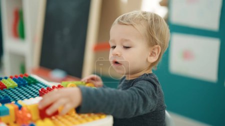 Foto de Adorable niño rubio jugando con bloques de construcción sentados en la mesa en el jardín de infantes - Imagen libre de derechos
