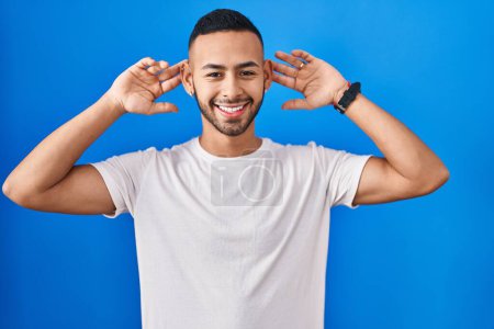 Foto de Young hispanic man standing over blue background smiling pulling ears with fingers, funny gesture. audition problem - Imagen libre de derechos