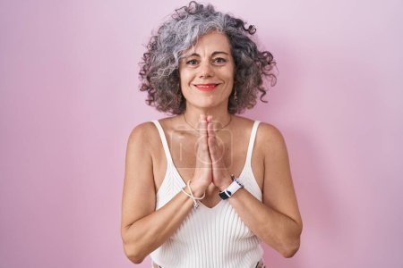 Foto de Mujer de mediana edad con el pelo gris de pie sobre fondo rosa rezando con las manos juntas pidiendo perdón sonriendo confiado. - Imagen libre de derechos