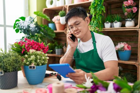 Photo for Young chinese man florist talking on smartphone using touchpad at flower shop - Royalty Free Image