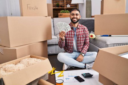 Téléchargez les photos : Middle east man with beard sitting on the floor at new home holding money looking positive and happy standing and smiling with a confident smile showing teeth - en image libre de droit