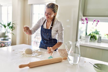 Foto de Joven mujer rubia sonriendo confiado hacer masa de pizza con las manos en la cocina - Imagen libre de derechos