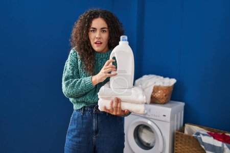 Foto de Mujer hispana joven sosteniendo la ropa y la botella de detergente expresión despistada y confusa. concepto de duda. - Imagen libre de derechos