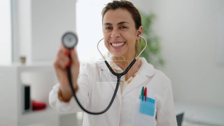Téléchargez les photos : Femme hispanique d'âge moyen portant un uniforme de médecin tenant un stéthoscope à la clinique - en image libre de droit