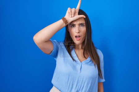 Photo for Young brunette woman standing over blue background making fun of people with fingers on forehead doing loser gesture mocking and insulting. - Royalty Free Image