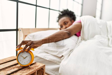 Foto de Mujer afroamericana apagando el despertador tumbado en la cama en el dormitorio - Imagen libre de derechos