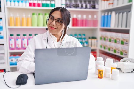 Téléchargez les photos : Jeune femme arabe travaillant à la pharmacie pharmacie en utilisant un ordinateur portable souriant regardant vers le côté et regardant loin en pensant. - en image libre de droit