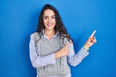 Téléchargez les photos : Jeune femme brune debout sur fond bleu souriant et regardant la caméra pointant avec deux mains et doigts sur le côté. - en image libre de droit