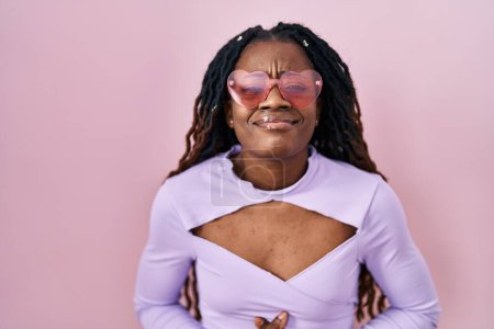 Téléchargez les photos : Femme africaine avec les cheveux tressés debout sur fond rose avec la main sur le ventre parce que les nausées, la maladie douloureuse se sentent mal. concept de douleur. - en image libre de droit