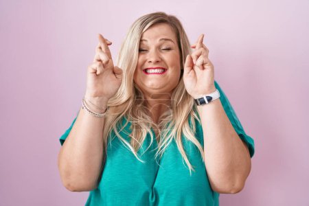 Téléchargez les photos : Caucasian plus size woman standing over pink background gesturing finger crossed smiling with hope and eyes closed. luck and superstitious concept. - en image libre de droit