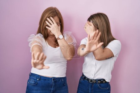 Téléchargez les photos : Hispanic mother and daughter wearing casual white t shirt over pink background covering eyes with hands and doing stop gesture with sad and fear expression. embarrassed and negative concept. - en image libre de droit