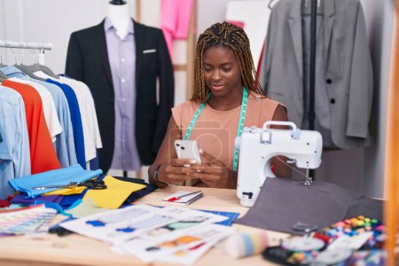 Téléchargez les photos : Femme afro-américaine tailleur souriant confiant en utilisant un smartphone au magasin de tailleur - en image libre de droit