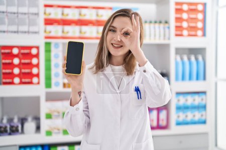 Foto de Mujer rubia caucásica que trabaja en la farmacia mostrando la pantalla del teléfono inteligente sonriendo feliz haciendo signo bien con la mano en el ojo mirando a través de los dedos - Imagen libre de derechos